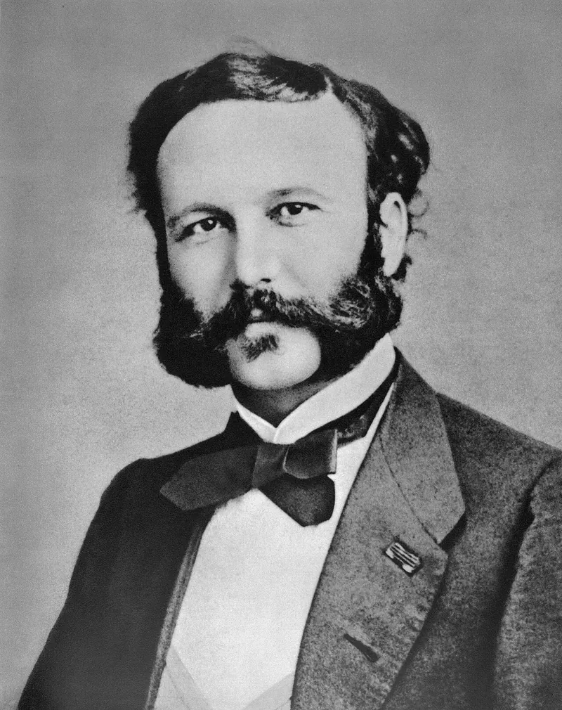 Black-and-white photographic portrait of a young Henry Dunant with dark hair and side whiskers, wearing a dark coat and cravat.